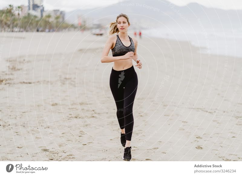 Female runner in stylish sportswear jogging along calm sandy seaside woman beach jumping summer healthy working out exercise female ocean activity training