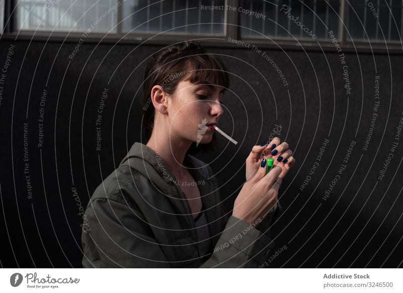 Female teenager in casual clothing smoking cigarette at city street light up holding liter wear jacket outfit grey wall nail blue brunette rebel sad modern