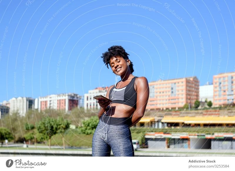 Cheerful African American sportswoman listening to music on smartphone listening music city happy smile sportive earphones mobile river bank jogger runner break
