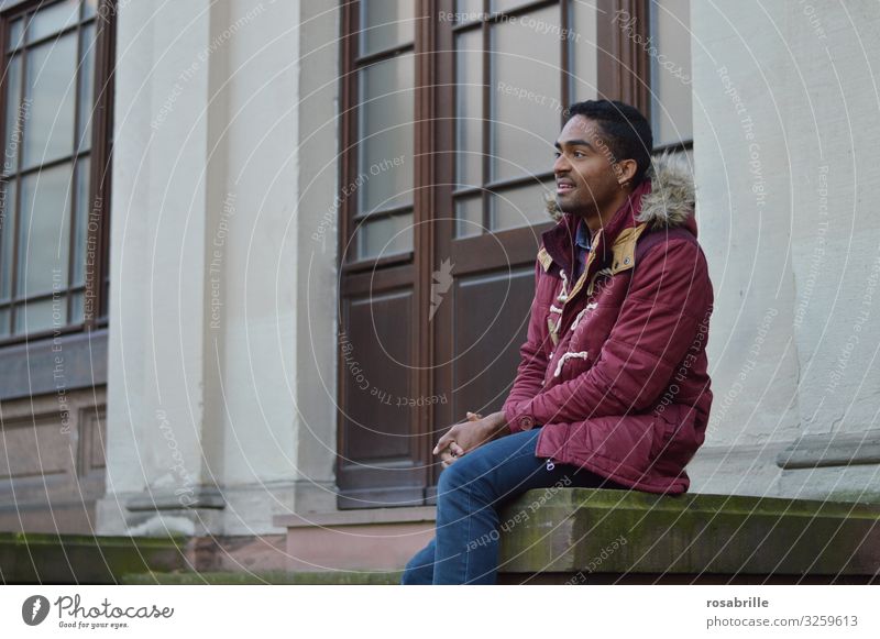 young man in red jacket Caucasian-African sits expectantly in front of a building Man youthful multiracial full of expectation delighted View into the distance