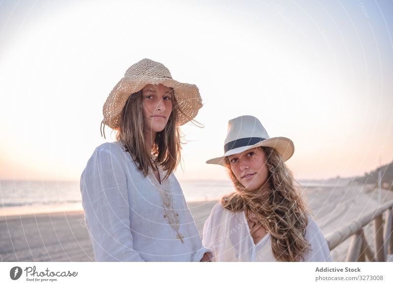 Smiling girlfriends in hats talking sitting on wooden fence on beach women sea traveling leaning seaside charming vacation curly holiday together freedom