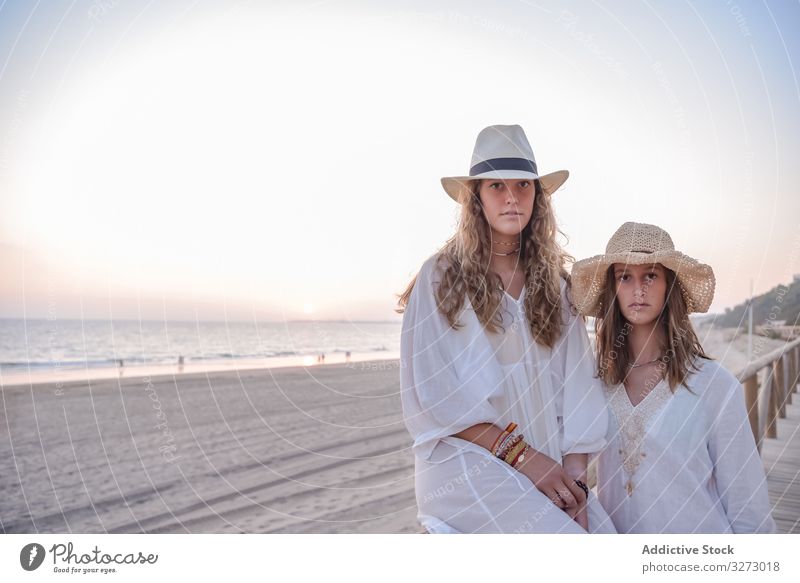 Smiling girlfriends in hats talking sitting on wooden fence on beach women speaking sea traveling leaning seaside charming vacation curly holiday together