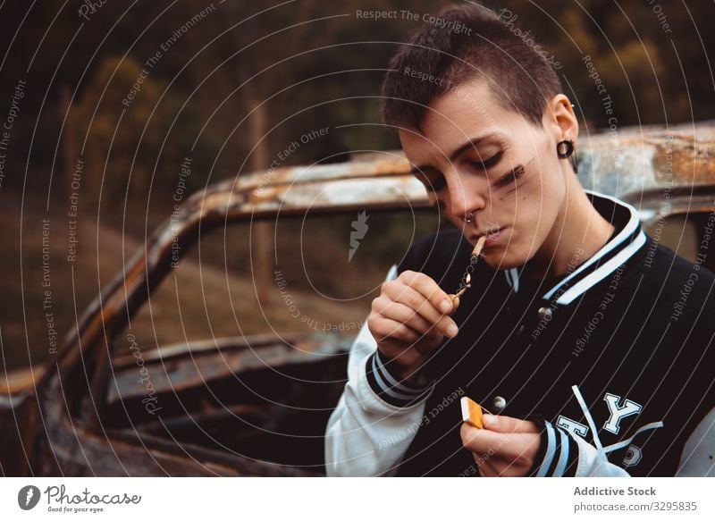 Young woman igniting cigarette near burnt car smoke rebel ignite rusty countryside lighter young lean lifestyle freedom vehicle auto androgynous short hair