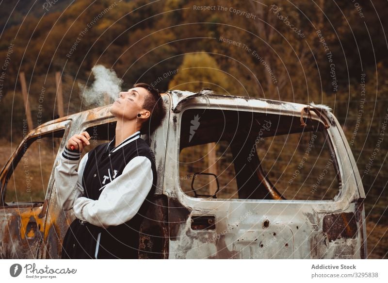 Young female smoking near damaged car woman cigarette smoke exhale rebel rusty countryside burnt young lean freedom vehicle auto androgynous short hair
