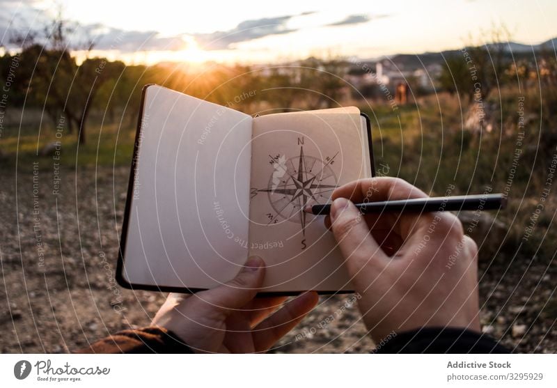 Crop traveler drawing compass during sunset countryside sketch notebook direction evening trip vacation weekend adventure guidance navigation lifestyle