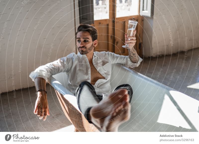 Serious cool barefoot rebel in elegant clothes relaxing alone in bathtub and celebrating own success against rustic interior in country house man alcohol eyes