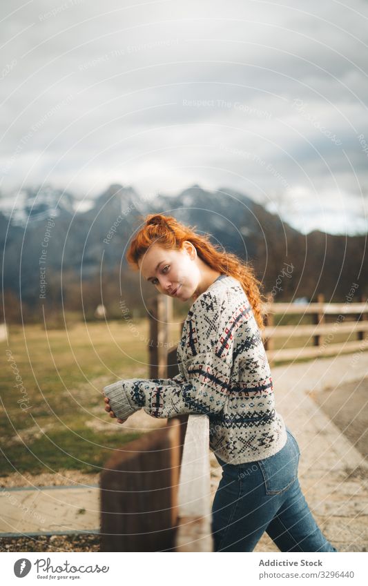 Red haired woman looking at camera leaning on wooden fence along road in mountains cozy countryside warm sweater comfortable fashion knitted relaxation human