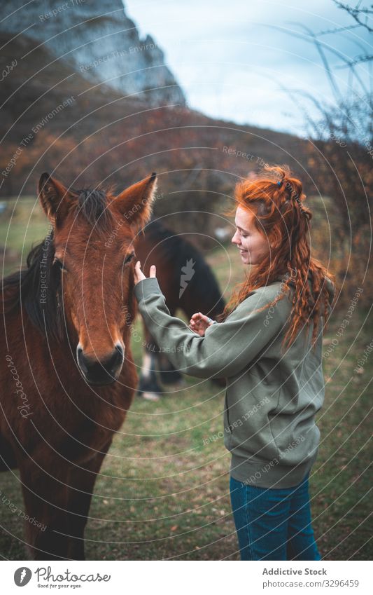 Red haired woman stroking horse in meadow animal care female mountain pasture grazing grass nature green field rural herd mammal fauna domestic red hair natural