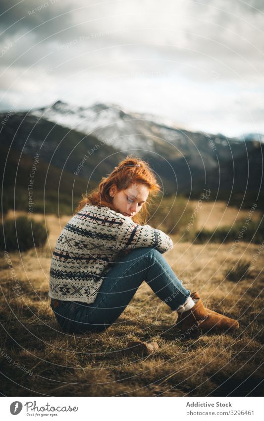 Red haired woman thoughtfully sitting on meadow in mountains cozy countryside pensive warm sweater comfortable fashion knitted relaxation human cheerful holiday