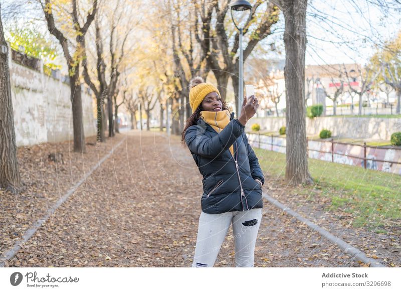 Smiling stylish black woman in hat and jacket taking selfie on smartphone in alley in park leaves female road using gadget nature african american natural