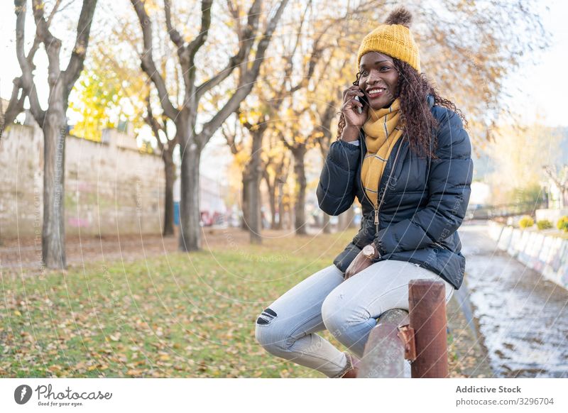 Joyful stylish black woman in hat and jacket speaking on smartphone in park leaves female road alley using gadget nature conversation african american talking