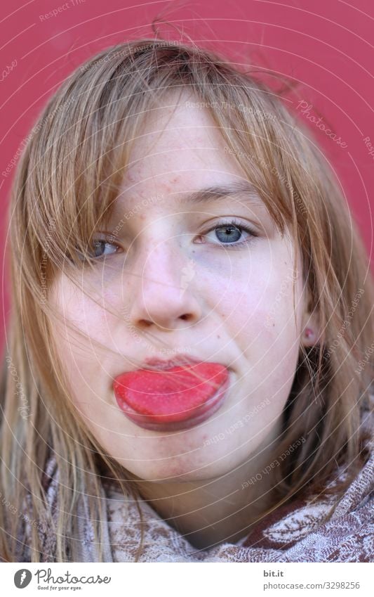 Girl, young woman eyes, red tongue sticking out. Tongue is stained red by food, paint, candy, berries or other food. In front of a red wall in the background.