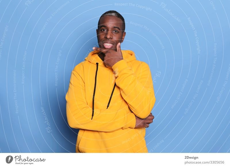 Studio shot of serious black man keeps hand under chin, wearing casual sweater, keeps hands partly crossed, poses against blue background, thinks about something