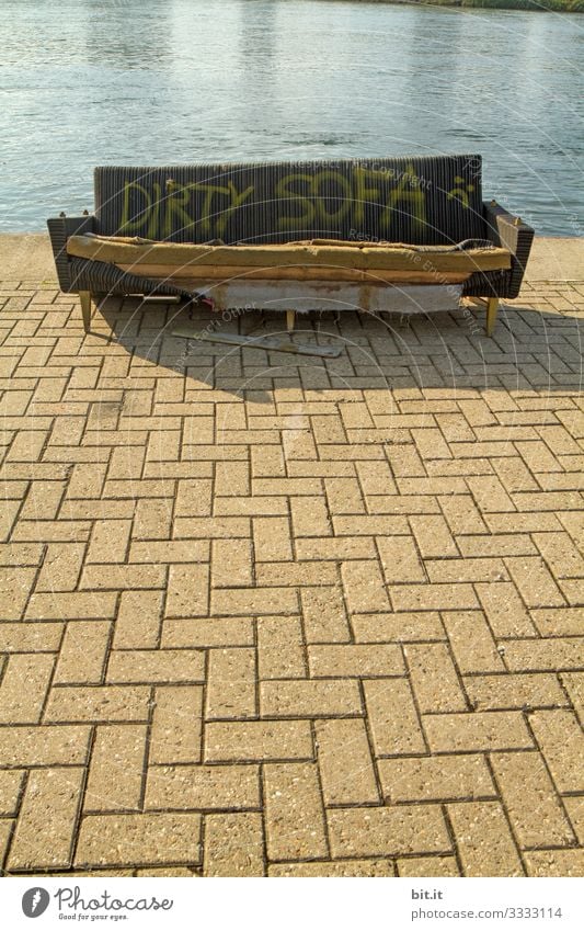 Old, broken, destroyed, brown, antique, dirty sofa with writing: Dirty Sofa, stands as a sign, symbol on a square with stone floor, at the water. Labeled couch in a beach bar, lounge stands to chill, sit, lie on the shore.