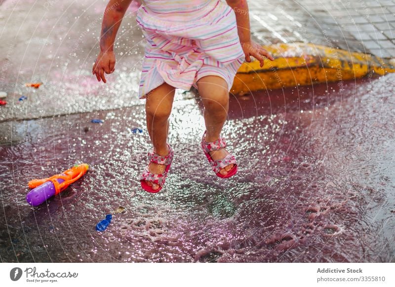 Crop girl jumping in colored water puddle festival paint dirty little street summer weekend kid child messy holiday celebration city town water gun toy play