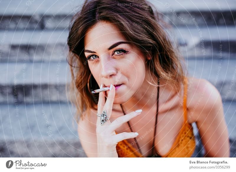 Beautiful young caucasian woman sitting on stairs at the city street on a sunny day and smoking a cigarette. Urban lifestyle and smoking concept. close up view