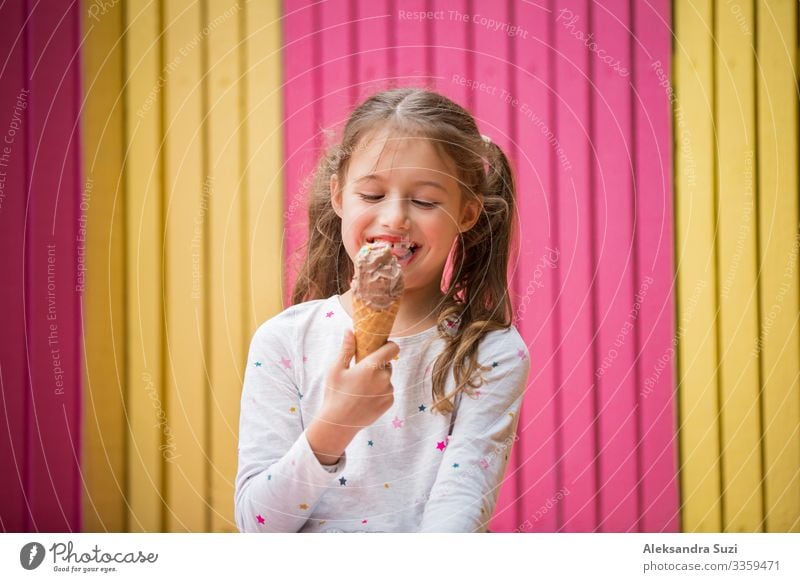 Cute little girl eating chocolate ice cream. Smiling and laughing. Colorful pink and yellow wall on background. Bright summer concept beautiful bright cafe
