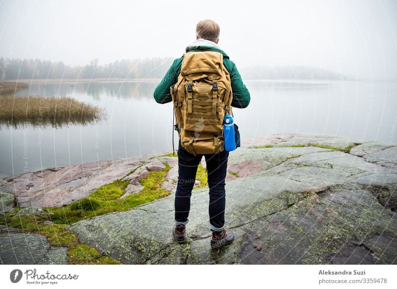 Mature man exploring Finland in the fall, looking into fog. Hiker with big backpack standing on mossy rock. Scandinavian landscape with misty sea and autumn forest. Back view