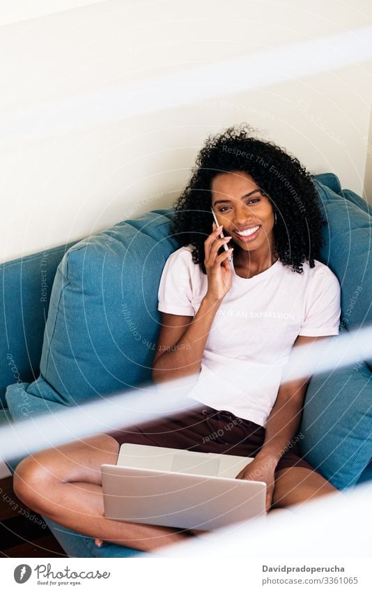 Content African American lady using devices at home woman laptop armchair freelance phone call smartphone browsing cozy comfort chat talk smile laugh enjoy