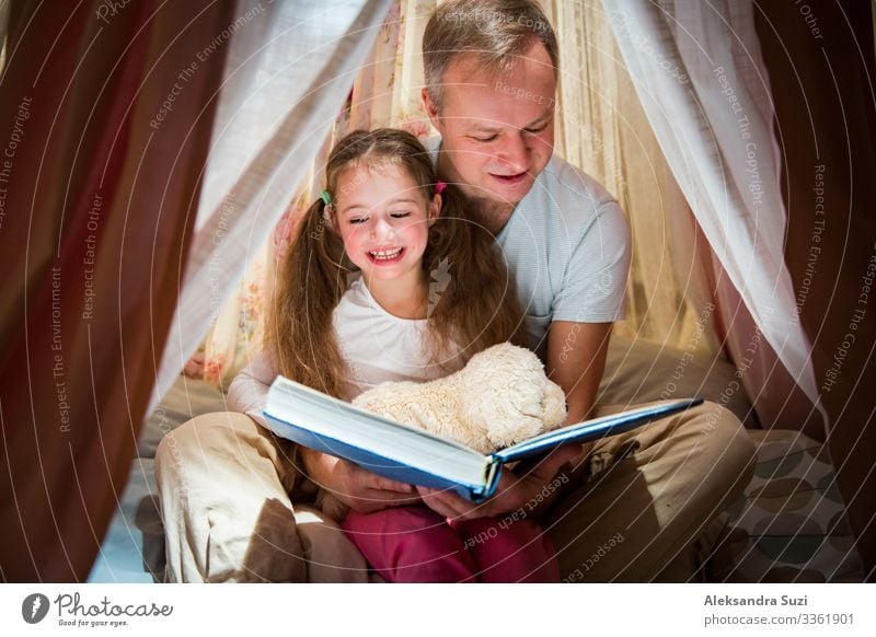 Family quality time. Father and daughter sit in homemade pink tent with flowers, read big book, look at each other, smile and laugh. Cozy stylish room. Family bonds concept
