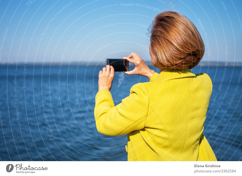 Woman in a yellow coat photographs the sea using a smartphone Lifestyle Style Beautiful Vacation & Travel Tourism Trip Sun Ocean PDA Screen Camera Adults