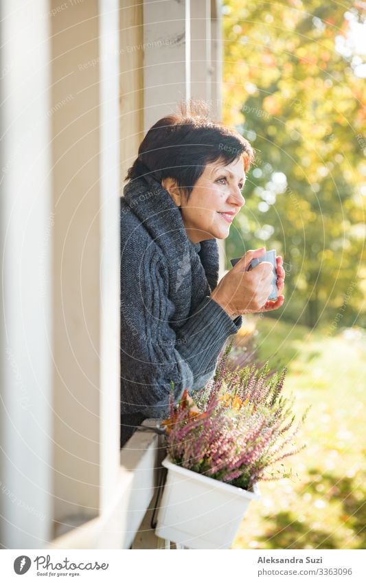 Mature attractive woman standing on Cozy wooden terrace with cup of hot coffee wrapped up in knitted warm sweater, happy smile. Lake house in autumn, yellow and red leaves on trees. Sunny day