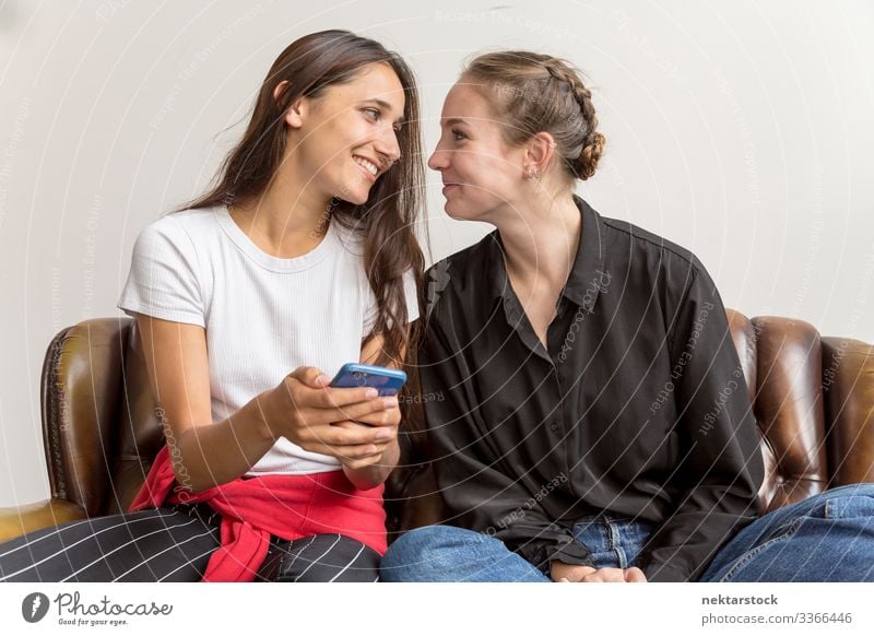 Two Women Seated Side by Side on Sofa Making Eye Contact smile smartphone eye contact eye to eye happiness looking at each other side view female girl