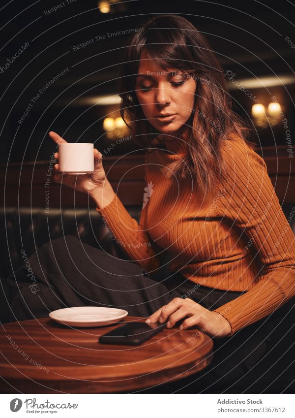 Pensive woman drinking coffee and browsing smartphone at table comfortable using gadget device stylish leather elegant thoughtful vogue lady trendy female