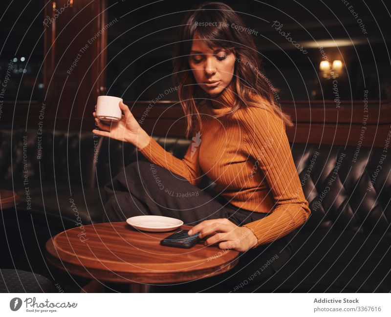 Pensive woman drinking coffee and browsing smartphone at table comfortable using gadget device stylish leather elegant thoughtful vogue lady trendy female