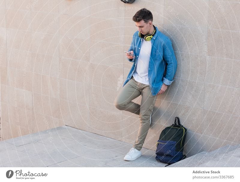 Student in headphones browsing smartphone leaning on wall student backpack using education man gadget campus building college surfing youth recreation device