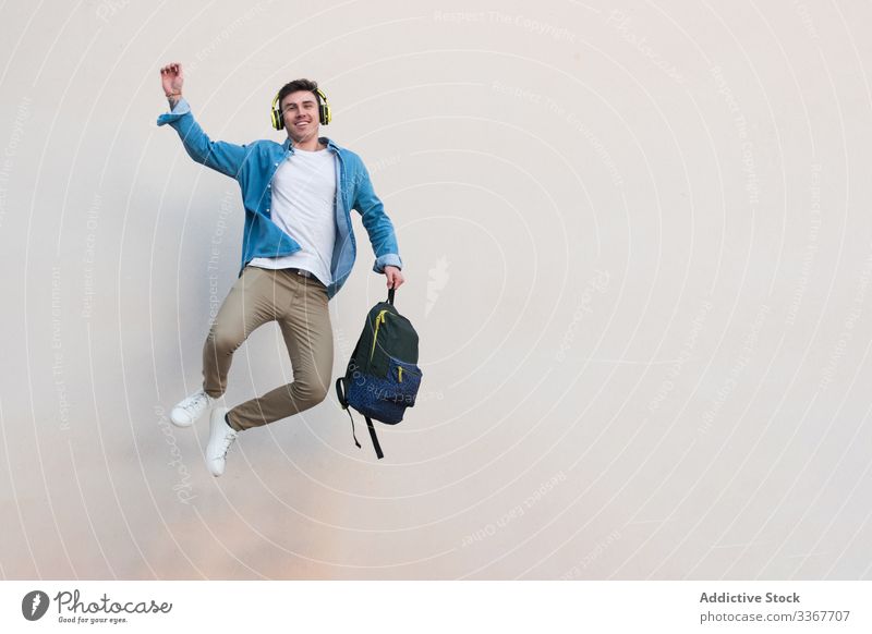 Joyful student in headphones with backpack jumping on background of wall victory education celebrating raising hand man building college listening youth smart