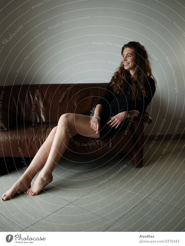Portrait of a young, tall woman sitting barefoot on a brown leather couch and smiling Style Joy already Well-being Flat (apartment) Sofa Young woman