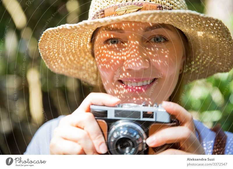 Close-up portrait of a beautiful woman in straw hat travelig in tropic forest, taking photos on retro camera. Light shadows through the cut-out detailed brim on face. Tourist with backpack.
