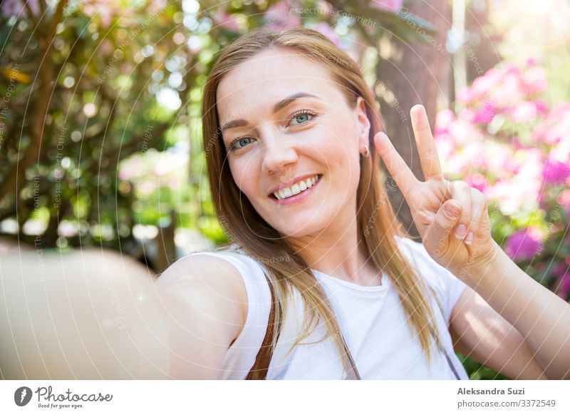 Beautiful woman traveling in tropic forest with flowers, walking along wooden path, taking selfie photos, smiling happily. Tourist with backpack showing peace sign