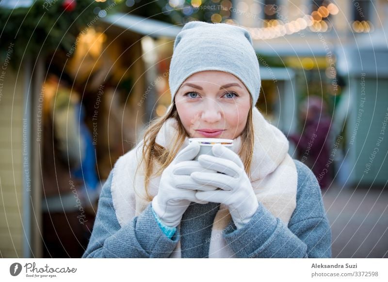 Young woman in Christmas market drinking cup of hot chocolate with marshmallow wearing knitted warm hat and scarf. Illuminated and decorated fair kiosks and shops on background. Helsinki, Finland