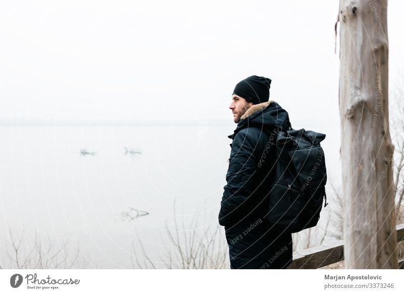 Young man standing by the river in winter morning active activity adult adventure alone backpack casual coast cold countryside day enjoy explorer foggy frost