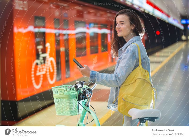 Teenager girl with backpack and bike standing on metro station holding smart phone in hand, scrolling and texting, smiling and laughing. Futuristic bright subway station. Finland, Espoo