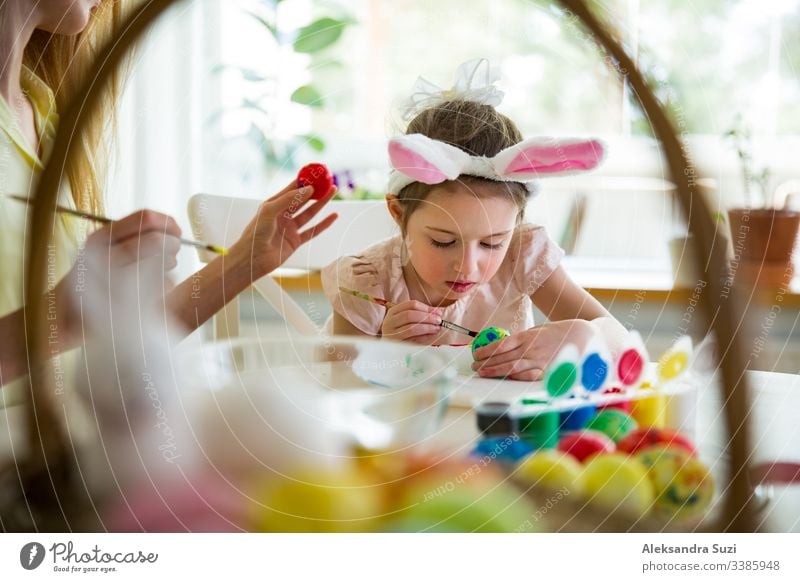 A mother and daughter celebrating Easter, painting eggs with brush. Happy family smiling and laughing. Cute little girl in bunny ears preparing the holiday.