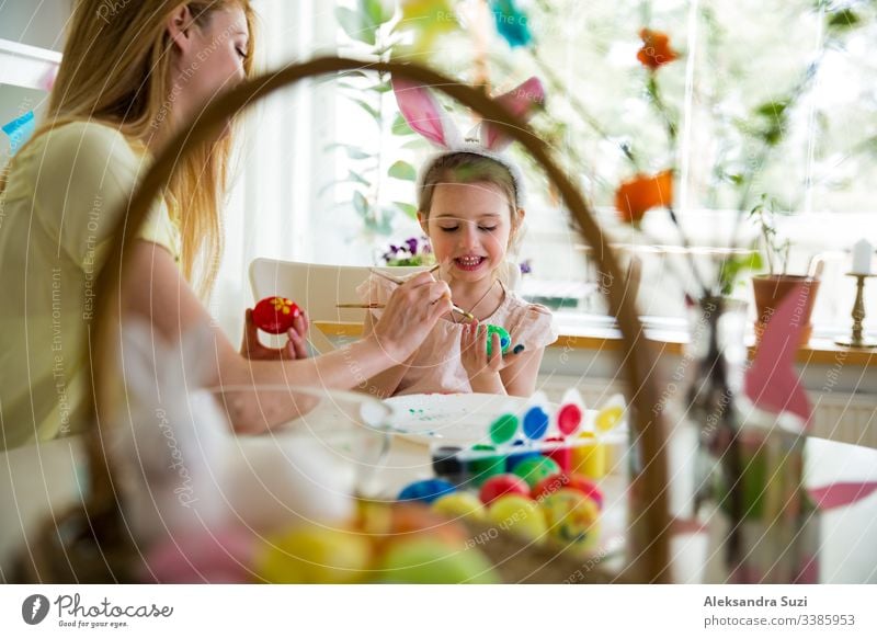 A mother and daughter celebrating Easter, painting eggs with brush. Happy family smiling and laughing. Cute little girl in bunny ears preparing the holiday.