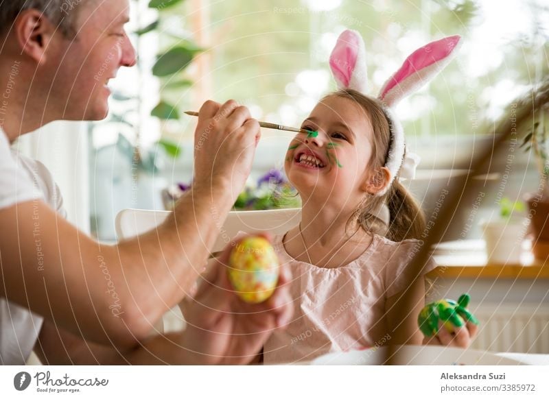 A father and daughter celebrating Easter, painting eggs with brush. Happy family smiling and laughing, drawing on face. Cute little girl in bunny ears preparing the holiday.