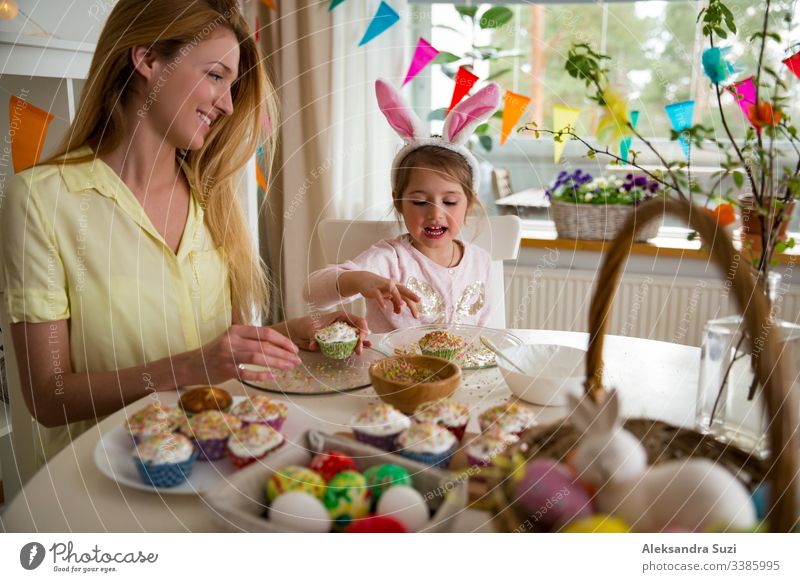 Mother and daughter celebrating Easter, cooking cupcakes, covering with glaze. Happy family holiday. Cute little girl in bunny ears. baking candid celebration