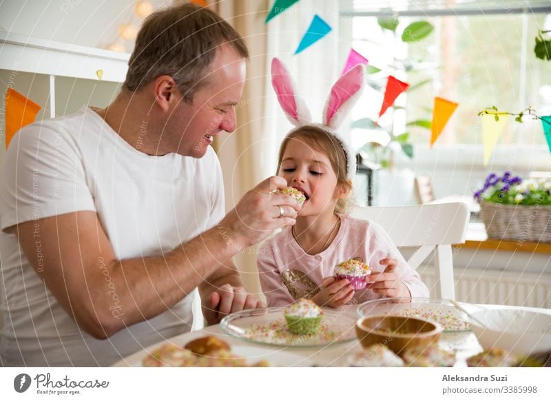 Father and daughter celebrating Easter, eating cupcakes covered with glaze. Happy family holiday. Cute little girl in bunny ears. Beautifully decorated room