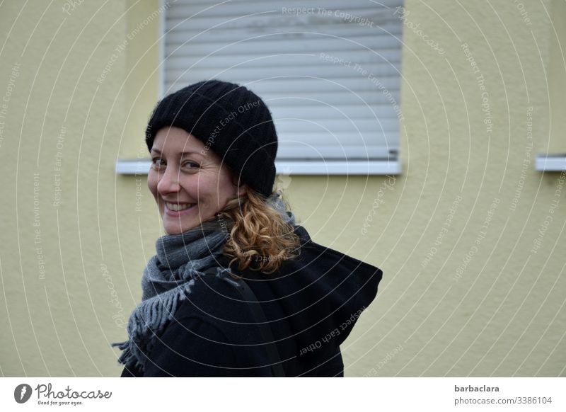 Life in front of a dreary wall roller shutter Woman Laughter House (Residential Structure) Curl cap Wall (building) Coat Black windowsill Gloomy Gray Autumnal