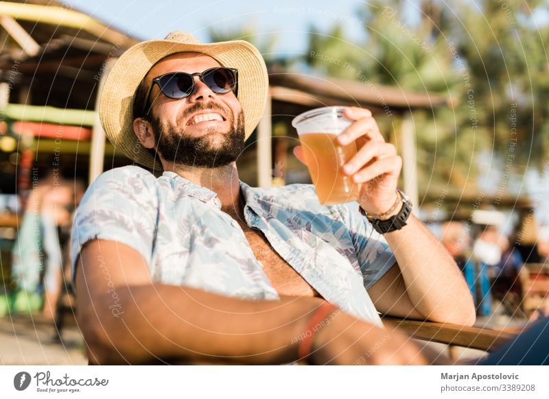 Young handsome man enjoying beer in beach bar 30s adult alcohol bearded beverage casual caucasian cheerful cheers cool destination drink fun happy hat hipster