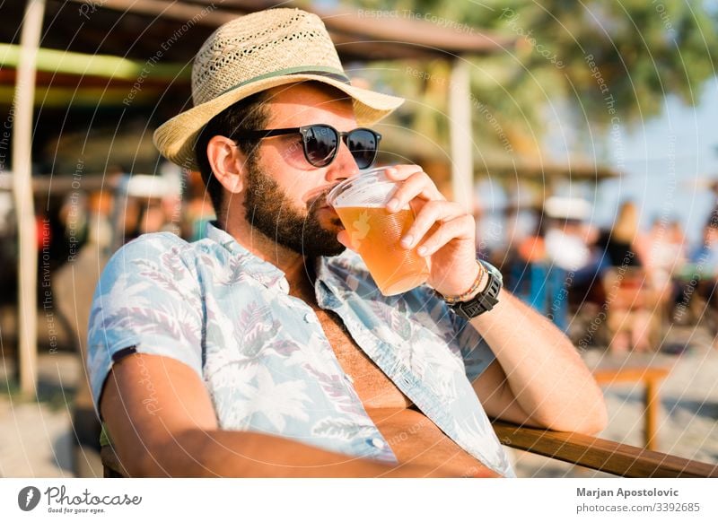 Young man drinking beer a beach bar enjoying sunset 30s adult alcohol alone bearded beverage casual caucasian cheers cool destination fun guy handsome hat