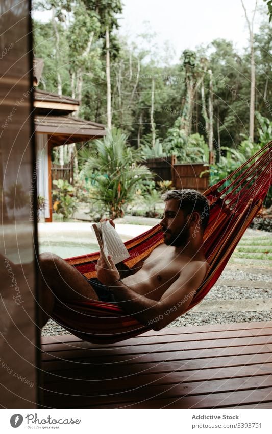 Young male tourist in swimwear with book on hammock man resort hotel lounge chill poolside read exotic tropical swimsuit relax rest sit lifestyle young enjoy