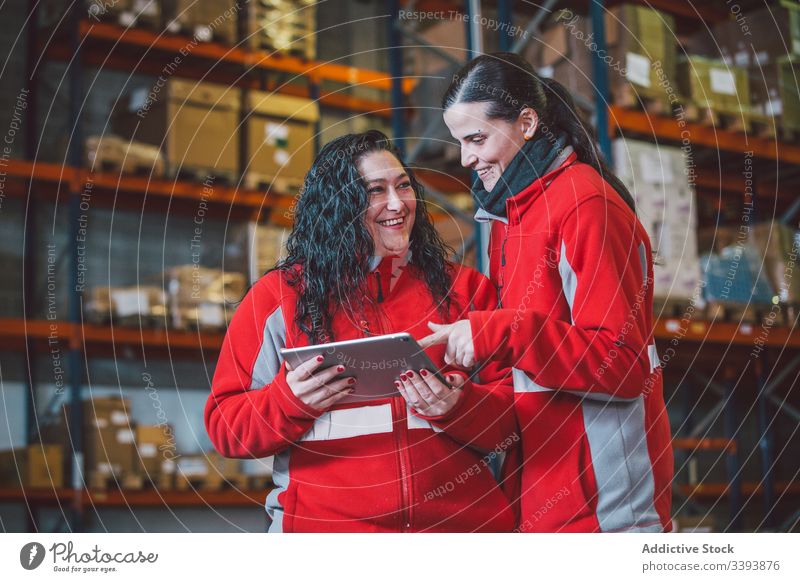 Satisfied smart female colleagues browsing digital tablet warehouse women using search watch device smile enjoy gadget storage logistic modern together work