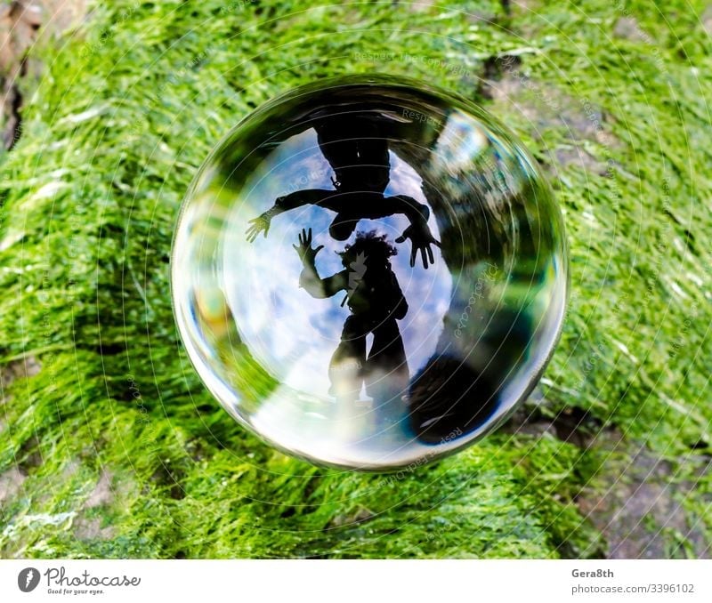 silhouettes of man and woman are distortedly reflected in glass ball lens lying on the green grass Man algae blue clouds curved mirror greeting hands pair palm