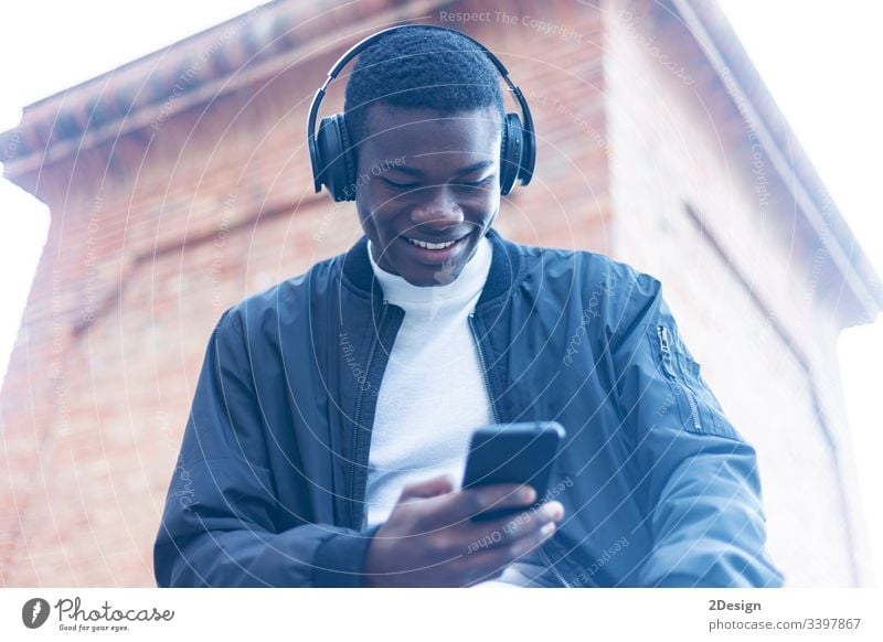 Young Black man standing in the street while listening music by headphones technology lifestyle male black casual attire trendy happy african earphones guy