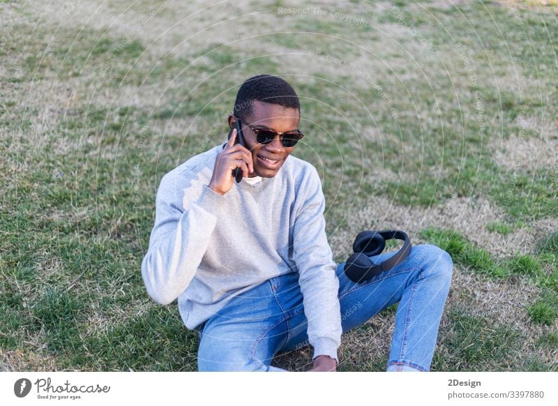 Fashionable black male with sunglasses sitting outdoors checking his news feed or messaging via social networks using cell phone outside lifestyle american